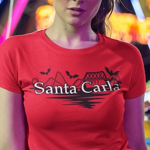 May include: A red t-shirt with the text "Santa Carla" in a stylized design. The shirt is worn in front of a brightly lit Ferris wheel at night. The design includes bats, a roller coaster, and palm trees.