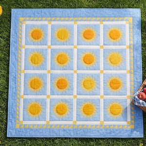 Può includere: Una trapunta quadrata caratterizzata da una griglia di sedici blocchi azzurri, ognuno centrato con un motivo a sole giallo. La trapunta è incorniciata da un bordo bianco e giallo con un motivo triangolare ripetuto e un bordo esterno azzurro.