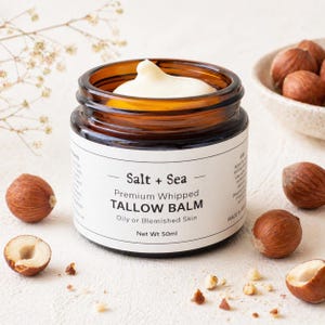 May include: An amber glass jar containing white balm, labelled "Salt + Sea Premium Whipped Tallow Balm" for oily or blemished skin. The jar is surrounded by whole and halved hazelnuts. The net weight of the balm is 50ml.