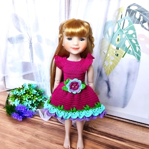 Puede incluir: Una muñeca con pelo largo y rojo claro que lleva un vestido de ganchillo magenta hecho a mano. El vestido presenta un cinturón floral verde y azul y un dobladillo festoneado con detalles verdes y azules. La muñeca está de pie frente a una ventana.