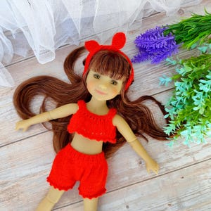 Könnte beinhalten: Ein rotes, gestricktes zweiteiliges Outfit für eine Puppe, bestehend aus einem Crop-Top und Shorts mit Häkelrand. Die Puppe hat langes braunes Haar und trägt ein rotes Stirnband mit Hasenohren.
