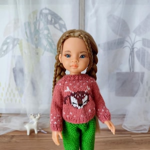 Può includere: Bambola con un maglione rosso lavorato a mano con un disegno di volpe bianca e marrone e pantaloni verdi. La bambola ha lunghi capelli castani intrecciati e occhi blu. Una piccola statuina di cervo bianco è sullo sfondo.