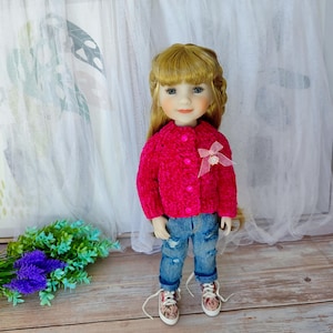 Może przedstawiać: Lalka ubrana w jaskraworóżowy, puszysty kardigan z różową kokardą, podarte niebieskie dżinsy i wzorzyste trampki. Lalka ma blond włosy i stoi przed białym zasłoną.