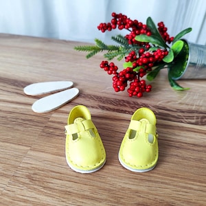 Può includere: Un paio di piccole scarpe in pelle gialla con cinturino e fibbia. Le scarpe hanno una suola bianca e sono appoggiate su una superficie di legno. Sono presenti anche due solette bianche. Un piccolo secchio di metallo con bacche rosse e foglie verdi è sullo sfondo.
