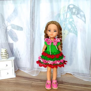 Könnte beinhalten: Puppe in einem handgefertigten grünen Häkelkleid mit roten und rosa Akzenten. Das Kleid hat einen Rüschenrock und eine rosa Schleife. Die Puppe hat lange, geflochtene braune Haare und trägt rosa Schuhe.