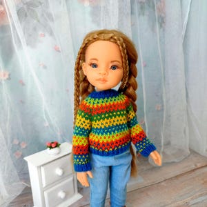 May include: A doll with long braided blonde hair, blue eyes, and a colorful striped sweater. The sweater features horizontal bands of red, orange, yellow, green, and blue. The doll is wearing blue jeans.