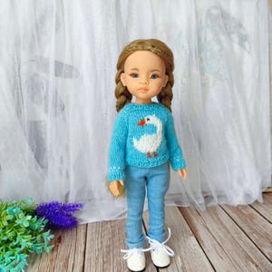 Op de afbeelding: Pop met een lichtblauwe gebreide trui met een witte ganzenontwerp en lichtblauwe jeans. De pop heeft blonde vlechten en witte schoenen met witte veters. De achtergrond omvat een wit gordijn en een houten oppervlak.