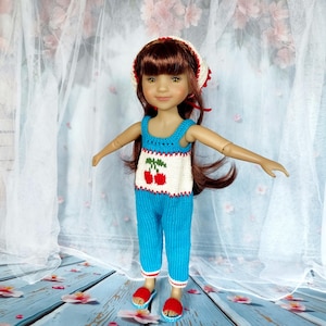 Può includere: Una bambola che indossa un completo in maglia con un motivo a ciliegie. L'outfit comprende una canotta e pantaloni blu, un cappello bianco con dettagli rossi e sandali rossi. La bambola ha i capelli castani ed è posizionata su uno sfondo azzurro.