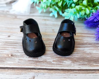 May include: A pair of miniature black Mary Jane shoes with a strap and buckle closure. The shoes have a textured sole and three small holes on the toe. The shoes are designed for dolls or other small figures.