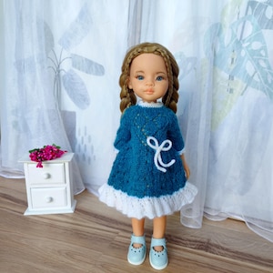 Puede incluir: Muñeca con un vestido de punto azul verdoso con ribete blanco y un lazo blanco. La muñeca tiene el pelo castaño trenzado y lleva zapatos azul claro. Una pequeña mesita de noche blanca con un ramo de flores rosas está en el fondo.