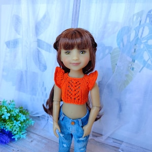 Könnte beinhalten: Eine Puppe trägt ein orangefarbenes, gestricktes Crop-Top und blaue Jeans. Das Oberteil hat ein detailliertes Lochmuster und Rüschenärmel. Die Puppe hat lange braune Haare und steht vor einem weißen Vorhang.