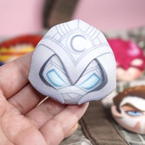 Hand holding a plush head pin badge button featuring a male character wearing a white suit with a hood and face bandages.