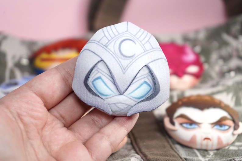 Hand holding a plush head pin badge button featuring a male character wearing a white suit with a hood and face bandages.