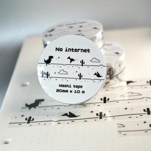 May include: A roll of white washi tape featuring a black pixelated dinosaur, cacti, clouds, and stars. The label reads No internet, Washi tape, and 20mm x 10m. The tape pattern is displayed on the surface below the roll.