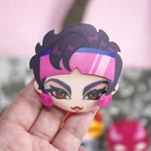 Hand holding a plush head pin badge button featuring a female character with brown eyes and hair as well as pink glasses and earrings.