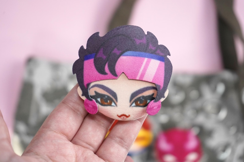 Hand holding a plush head pin badge button featuring a female character with brown eyes and hair as well as pink glasses and earrings.