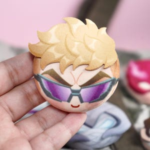 Hand holding a plush head pin badge button featuring a male character with blonde hair and purple googles.