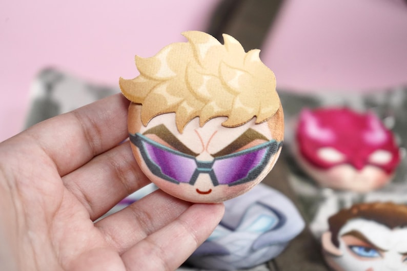 Hand holding a plush head pin badge button featuring a male character with blonde hair and purple googles.