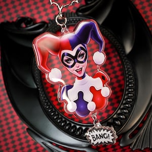 Puede incluir: Un llavero temático de Harley Quinn con una imagen de dibujos animados del personaje con su atuendo de arlequín rojo, blanco y azul. El llavero incluye un dije que dice "BANG!" y una cadena de metal plateado.