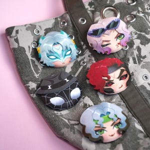 May include: Five circular pins featuring stylized anime-inspired character faces attached to a grey and white camouflage-patterned fabric bag. The pins display various designs including a masked figure, a character with sunglasses, a red-haired figure, a black spider-web patterned mask, and a character with green accents.