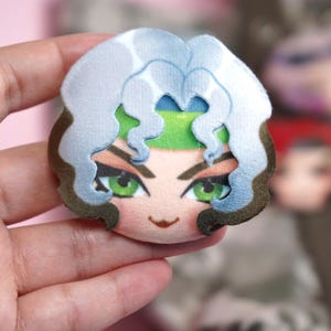 Hand holding a plush head pin badge button featuring a female character with green eyes as well as white and brown hair.