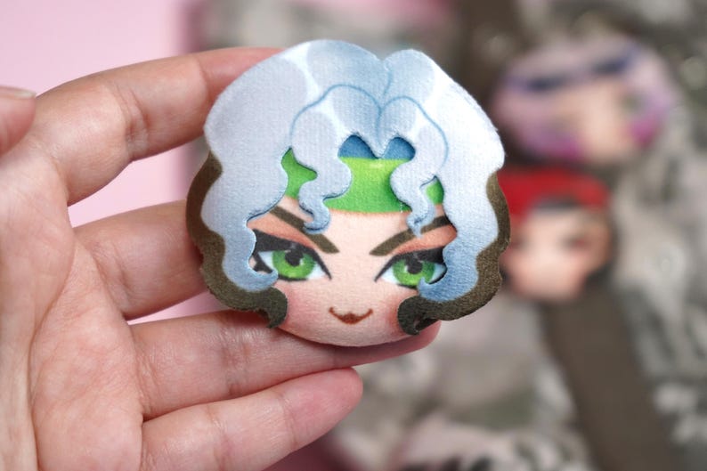 Hand holding a plush head pin badge button featuring a female character with green eyes as well as white and brown hair.