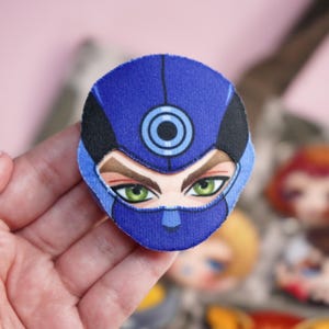 Hand holding a plush head pin badge button featuring a male character wearing a blue face mask with target symbol.