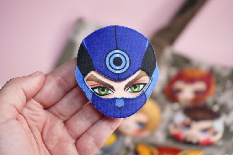 Hand holding a plush head pin badge button featuring a male character wearing a blue face mask with target symbol.