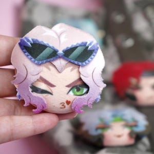 Hand holding a plush head pin badge button featuring a female character with green eyes, pink hair and purple sunglasses as well as a flirty kissing expression.