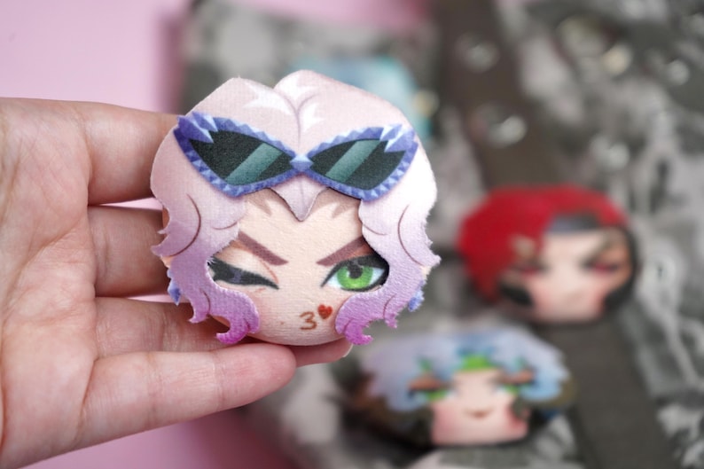 Hand holding a plush head pin badge button featuring a female character with green eyes, pink hair and purple sunglasses as well as a flirty kissing expression.