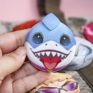Hand holding a plush head pin badge button featuring a blue shark character smiling with its tongue out.