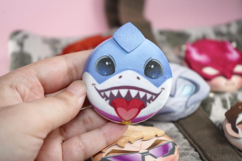 Hand holding a plush head pin badge button featuring a blue shark character smiling with its tongue out.