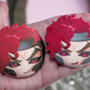 Hand holding two plush head pin badge buttons featuring a male character with pink eyes, red hair and a black helmet as well as a cheeky grin.
