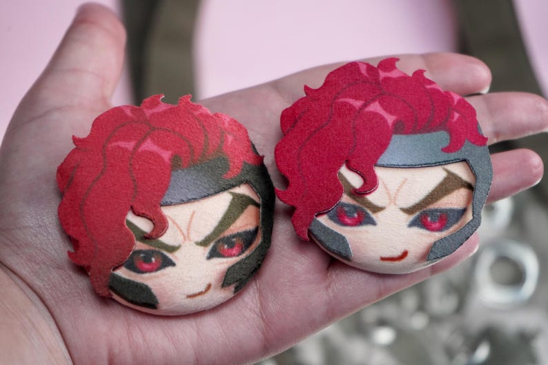 Hand holding two plush head pin badge buttons featuring a male character with pink eyes, red hair and a black helmet as well as a cheeky grin.
