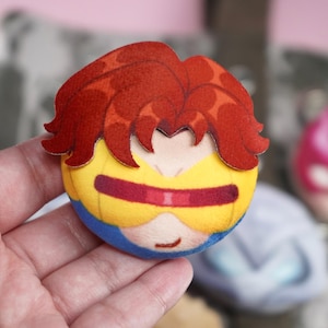 Hand holding a plush head pin badge button featuring a male character with orange hair and yellow googles.