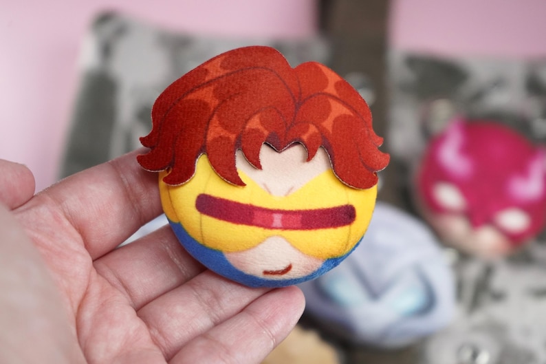 Hand holding a plush head pin badge button featuring a male character with orange hair and yellow googles.