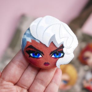 Hand holding a plush head pin badge button featuring a female character with dark complexion, blue eyes and white hair.