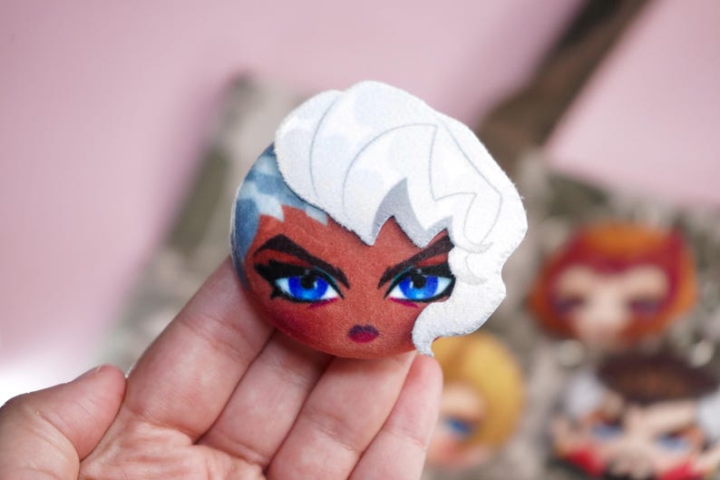 Hand holding a plush head pin badge button featuring a female character with dark complexion, blue eyes and white hair.