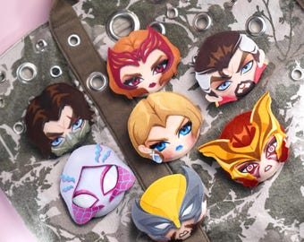 May include: A collection of seven embroidered character patches featuring stylized faces and masks, including figures with red, brown, blonde, and yellow hair, arranged on a grey and white camouflage-patterned fabric bag with metal grommets.