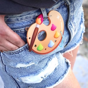 Op de afbeelding: Een beige, ovaalvormige kunstenaarspalet-bedel met een bruine kwast met roze punt, geaccentueerd door kleine kleurrijke verfspatten in roze, geel en blauw, bevestigd aan een blauwe metalen kogelketting en hangend aan een riemlus van een spijkerbroek.