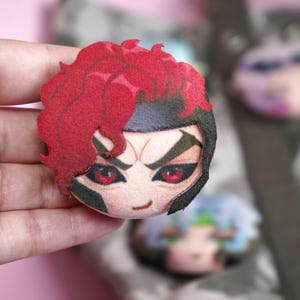 Hand holding a plush head pin badge button featuring a male character with pink eyes, red hair and a black helmet as well as a cheeky grin.