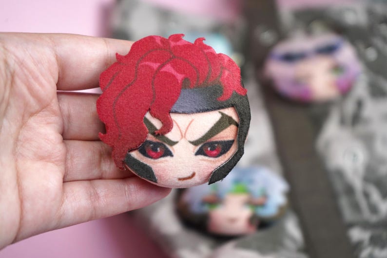 Hand holding a plush head pin badge button featuring a male character with pink eyes, red hair and a black helmet as well as a cheeky grin.