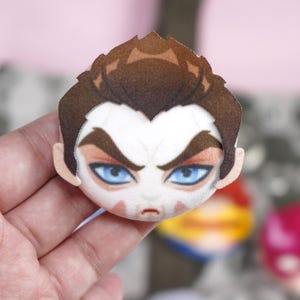 Hand holding a plush head pin badge button featuring a male character with blue eyes, brown hair and a white face paint shaped like a skull.