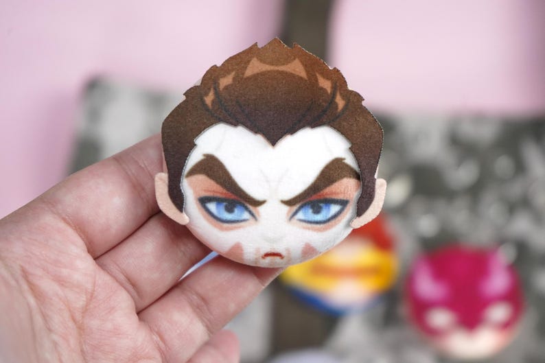 Hand holding a plush head pin badge button featuring a male character with blue eyes, brown hair and a white face paint shaped like a skull.