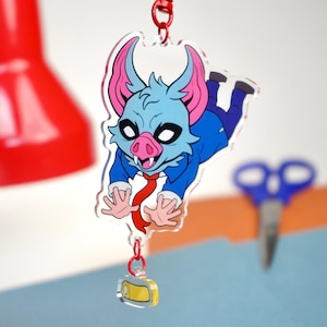 May include: A clear acrylic keychain featuring a cartoon bat-like creature with blue fur, pink ears, and a red tie. The keychain is suspended by a red chain and includes a small, rectangular charm with yellow and white details. The creature is wearing a blue suit.
