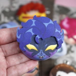 Hand holding a plush head pin badge button featuring a male character with yellow eyes, blue skin, pointy ears and face markings.