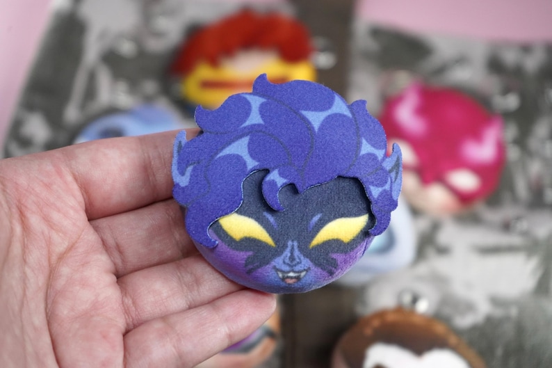 Hand holding a plush head pin badge button featuring a male character with yellow eyes, blue skin, pointy ears and face markings.