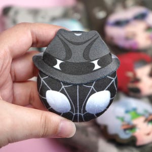 Hand holding a plush head pin badge button featuring a male character dressed in a black noir outfit and a hat.