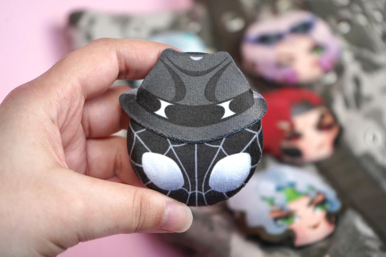 Hand holding a plush head pin badge button featuring a male character dressed in a black noir outfit and a hat.