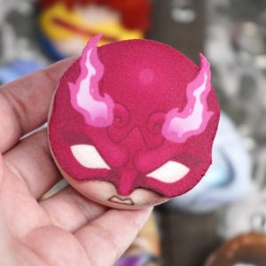 Hand holding a plush head pin badge button featuring a male character in a red face mask with horns.